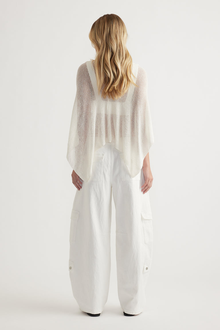 Woman viewed from behind wearing a sheer white asymmetric knit poncho with white cargo pants