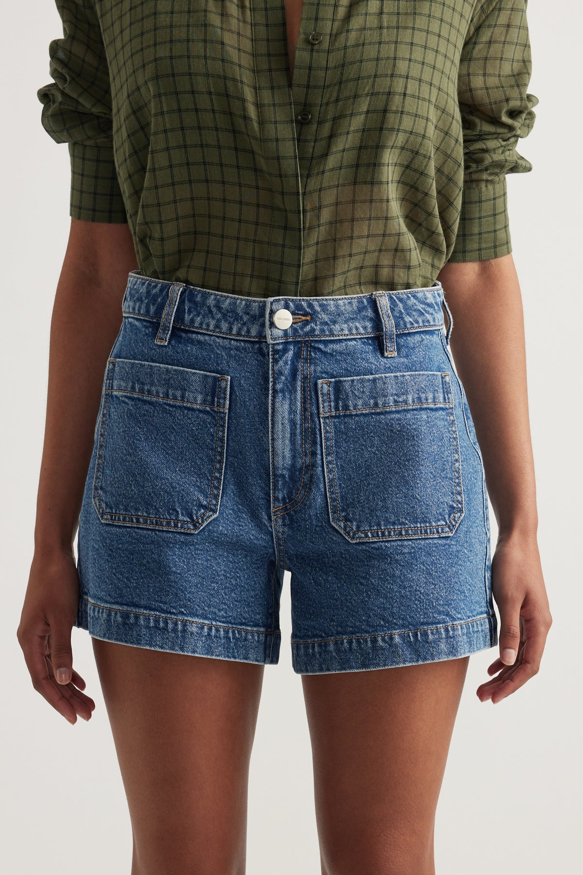 Woman wearing high rise blue denim shorts with front patch pockets and a rolled hem