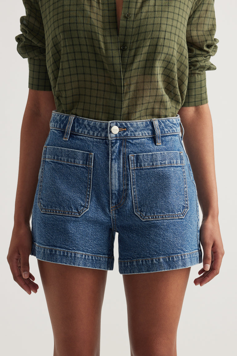Woman wearing high rise blue denim shorts with front patch pockets and a rolled hem