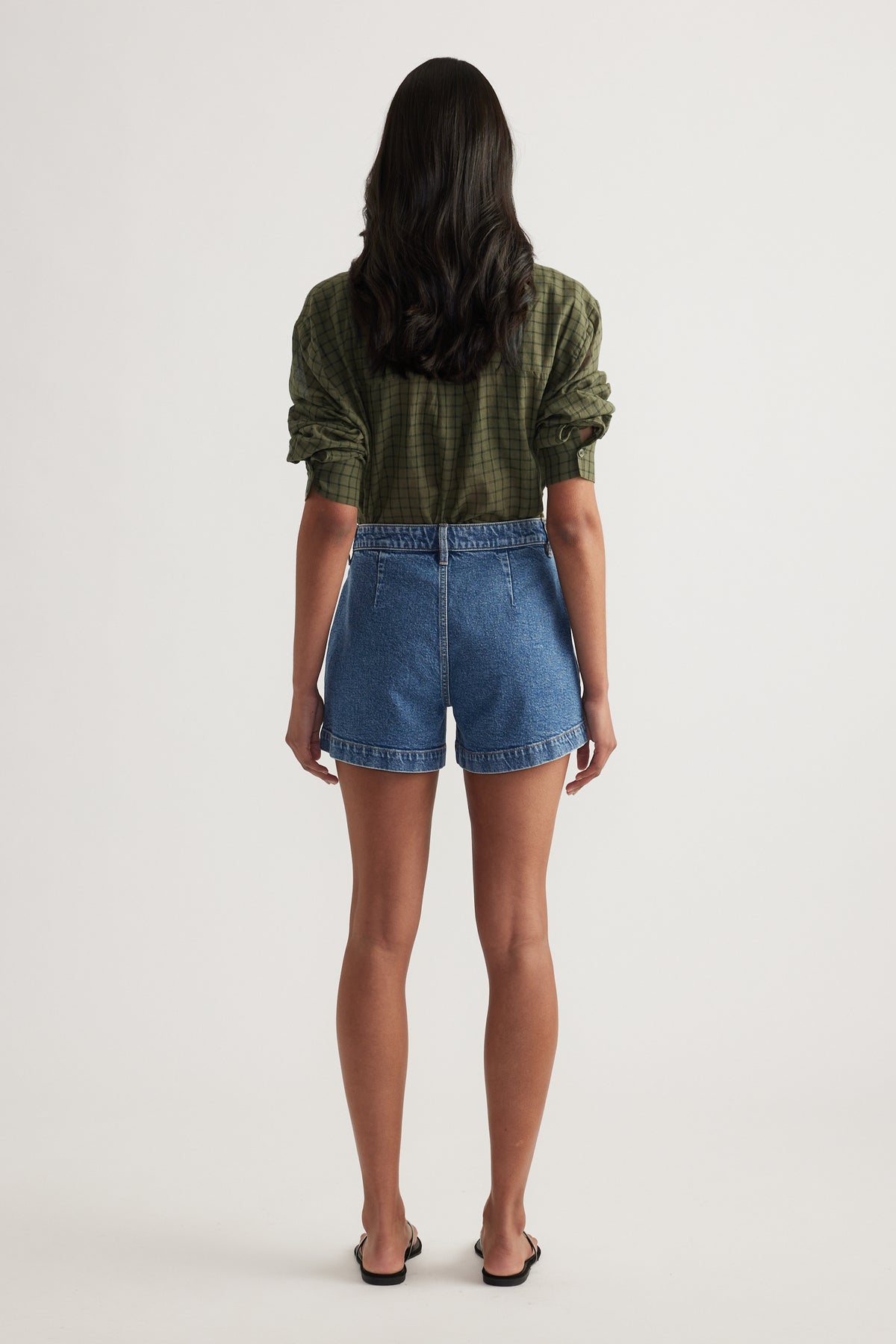 Back view of a woman wearing high rise blue denim shorts with rolled hems and a green plaid shirt