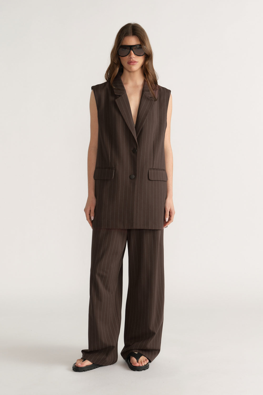 Woman wearing sleeveless dark brown pinstripe vest with matching wide-leg pants and black sandals on neutral background