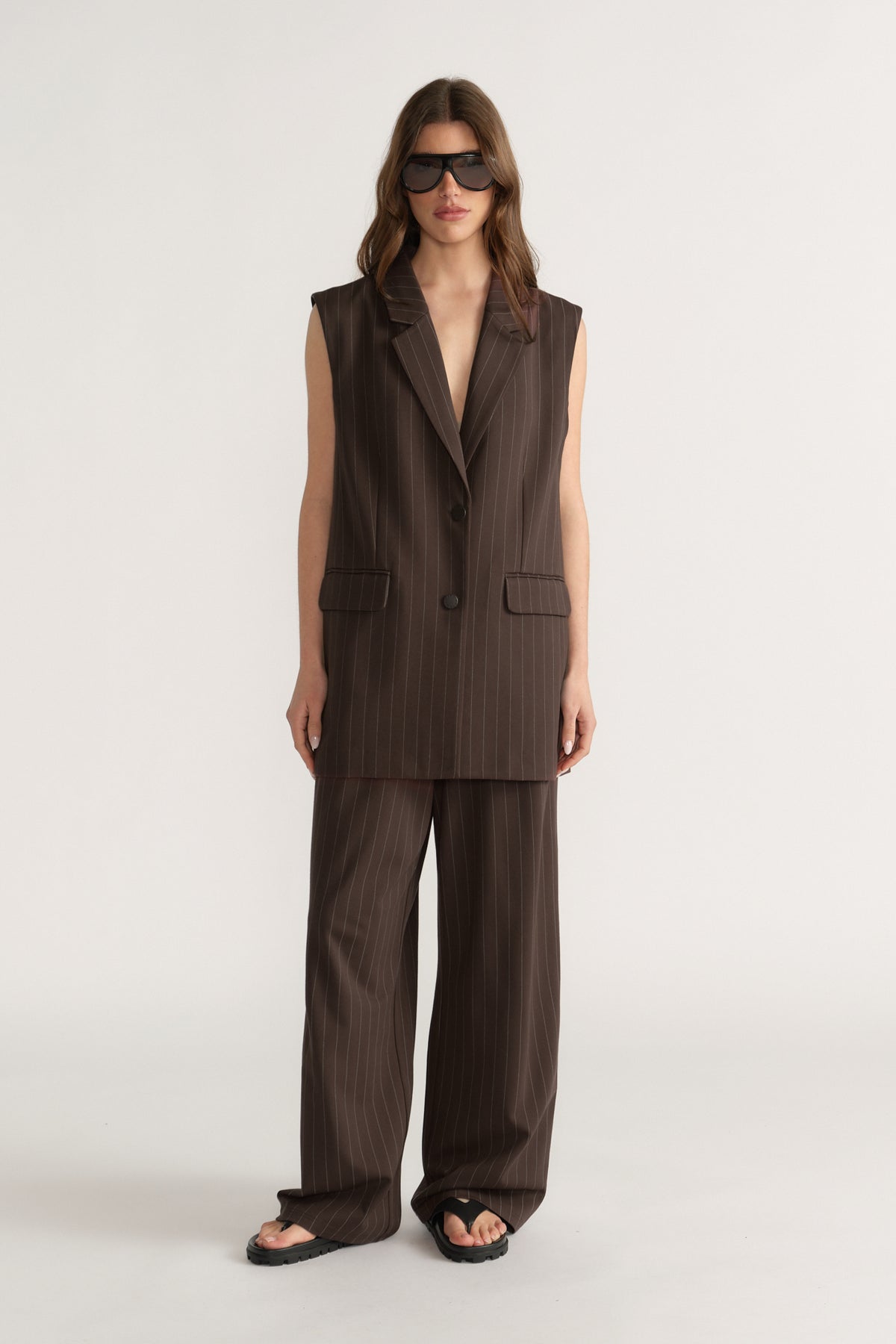 Woman wearing sleeveless dark brown pinstripe vest with matching wide-leg pants and black sandals on neutral background