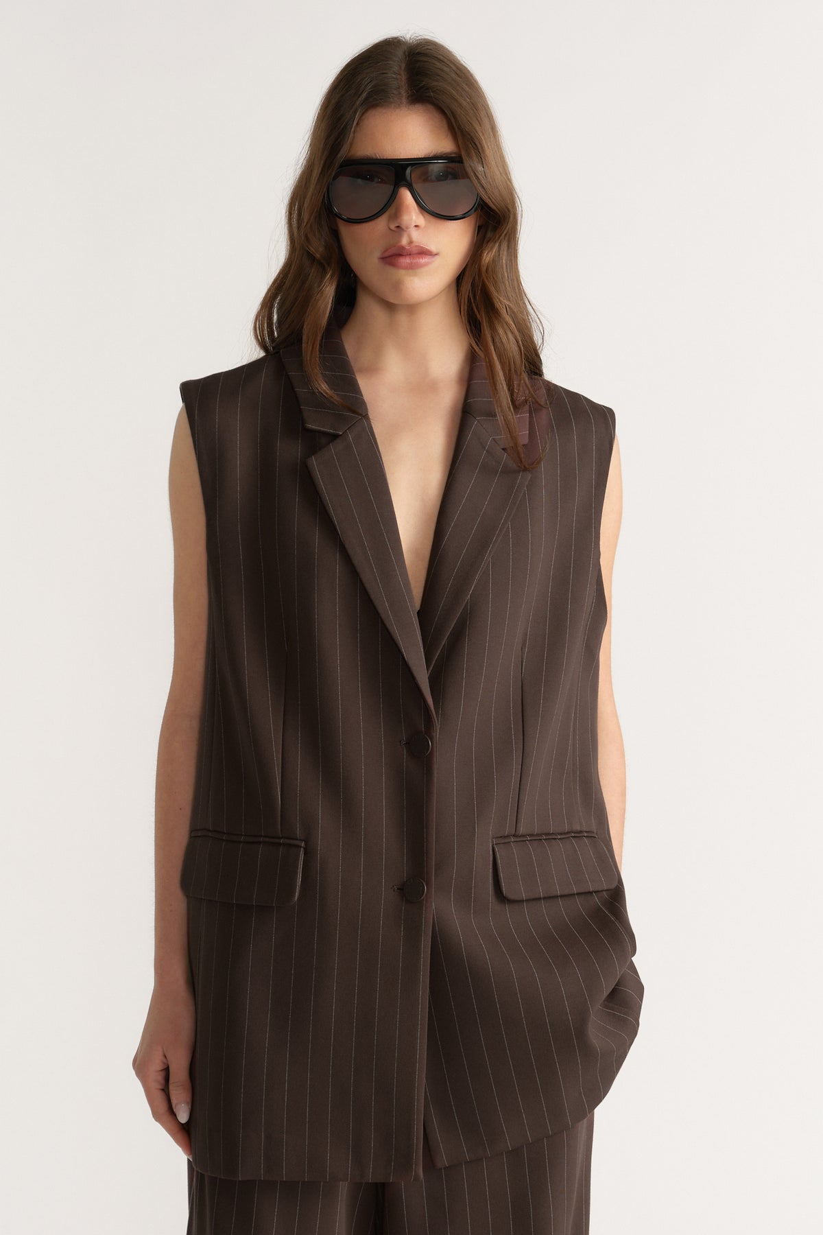 Woman wearing sleeveless dark brown pinstripe vest with padded shoulders and front pockets, styled with sunglasses