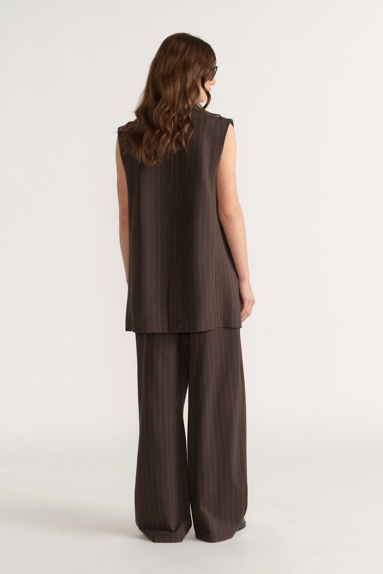 Woman wearing sleeveless dark brown pinstripe vest and matching wide-leg pants, standing against plain background