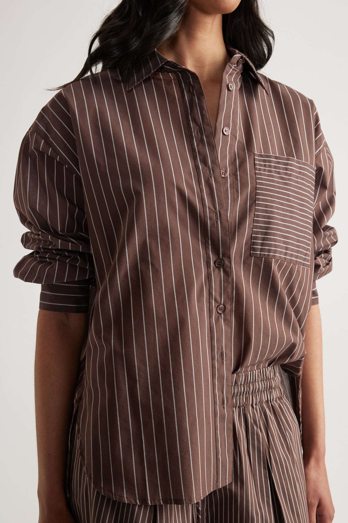 Woman wearing a brown oversized striped button-up shirt with rolled sleeves and matching pants