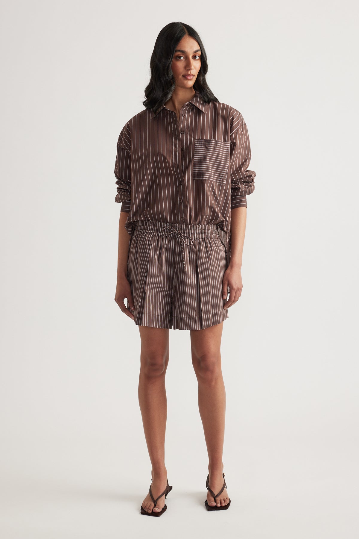 Woman wearing a brown striped button up shirt with matching pull on shorts and flat sandals