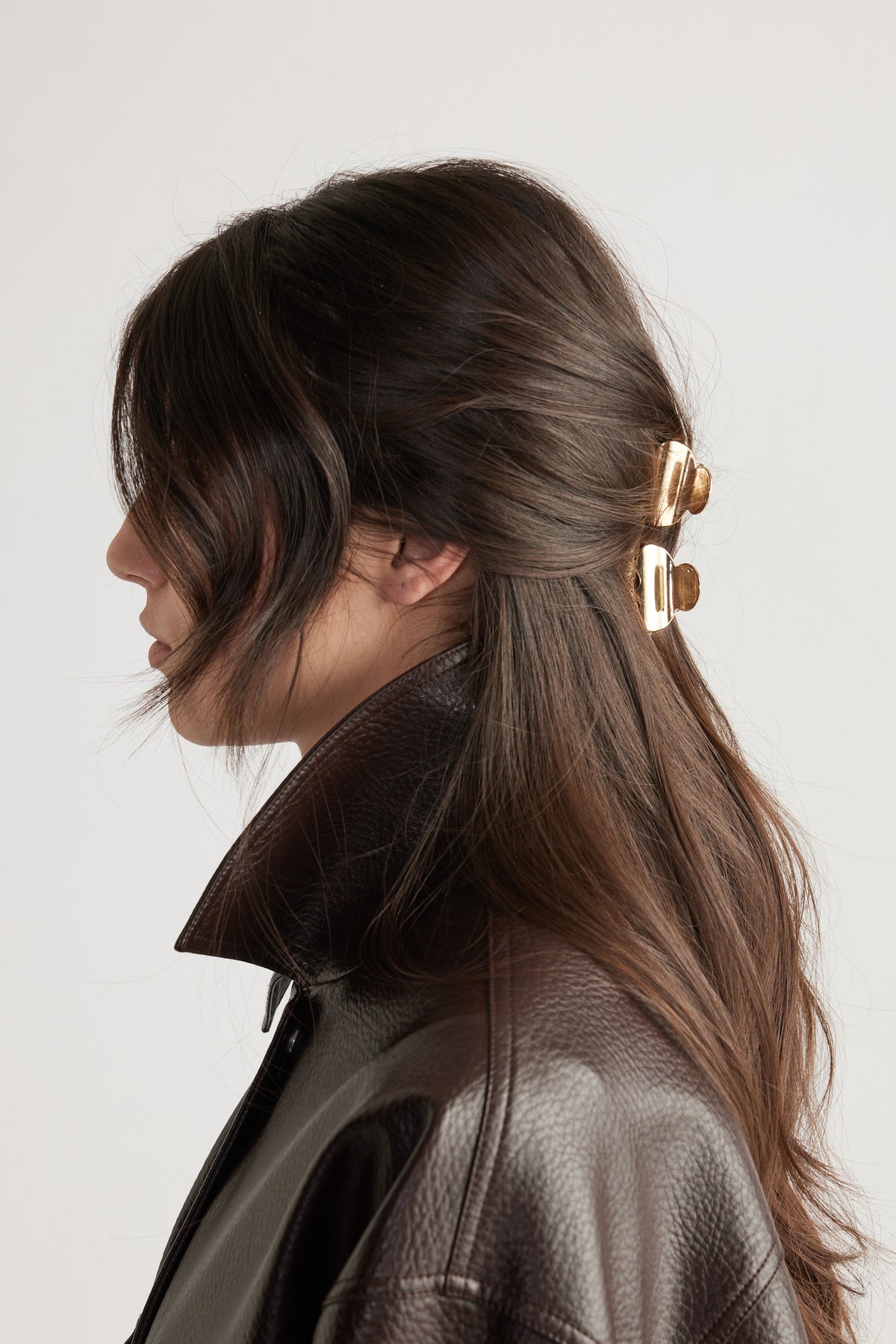 Profile of woman with dark brown hair styled using two gold mini hair claws, wearing a textured dark leather jacket with raised collar