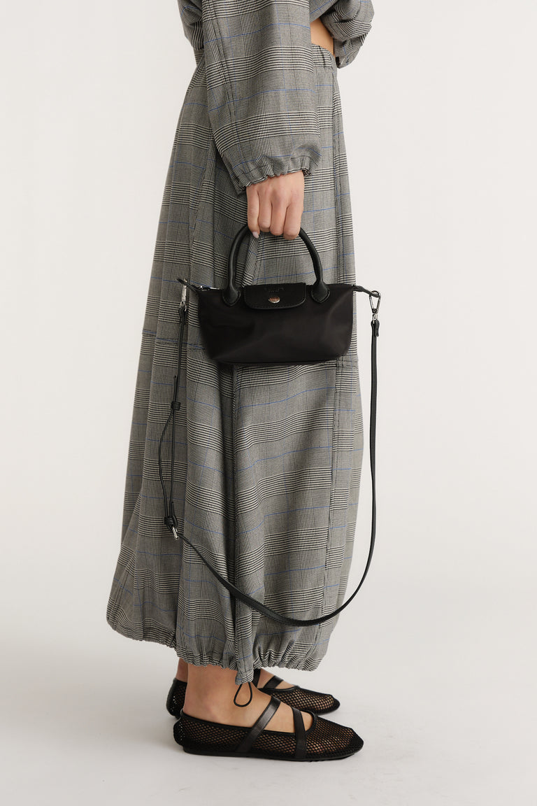 Woman holding a small black handbag with top handles and a long strap, styled with a plaid dress and black mesh flats.