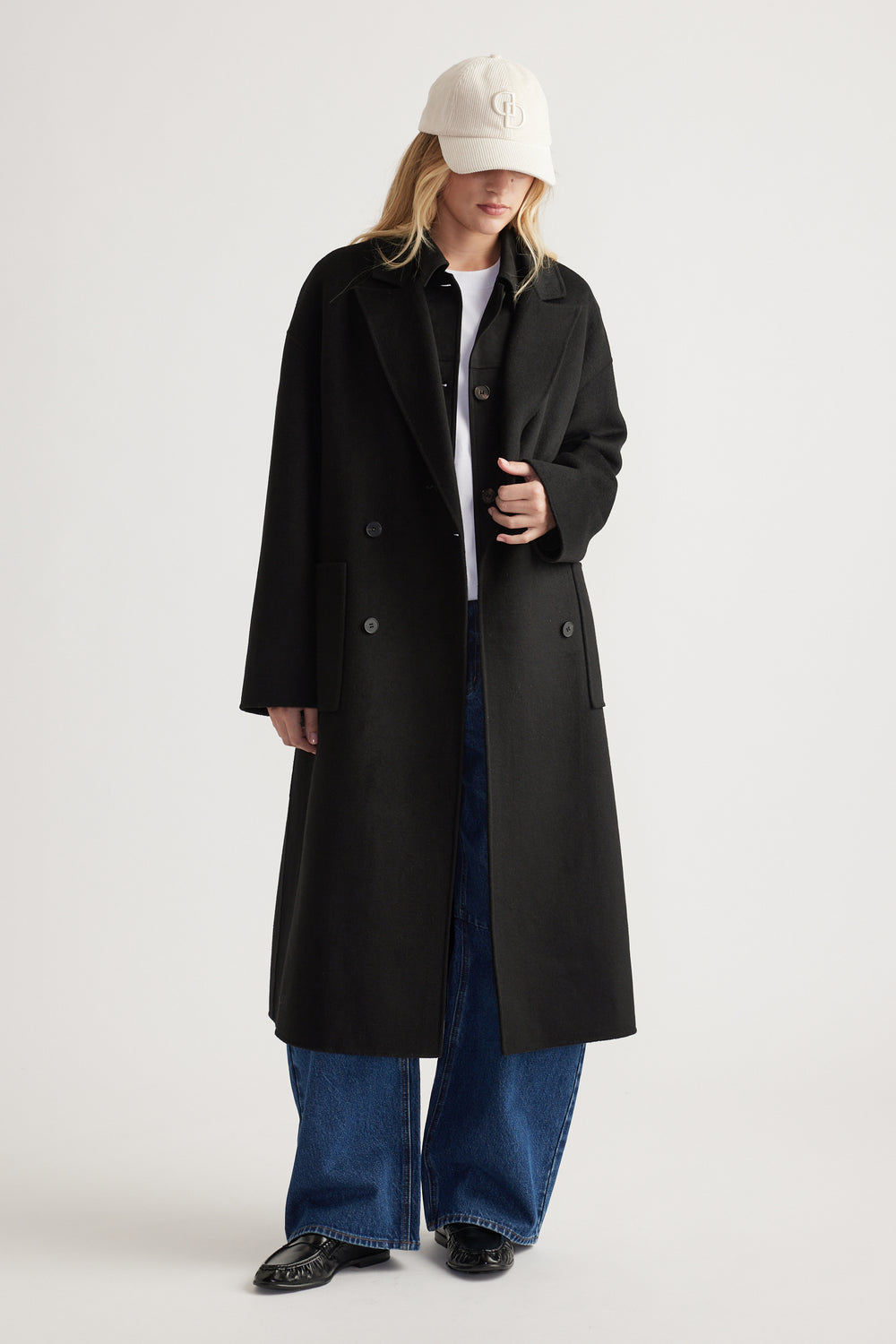 Woman wearing a long black coat over blue wide-leg jeans and black loafers, paired with a beige cord baseball cap