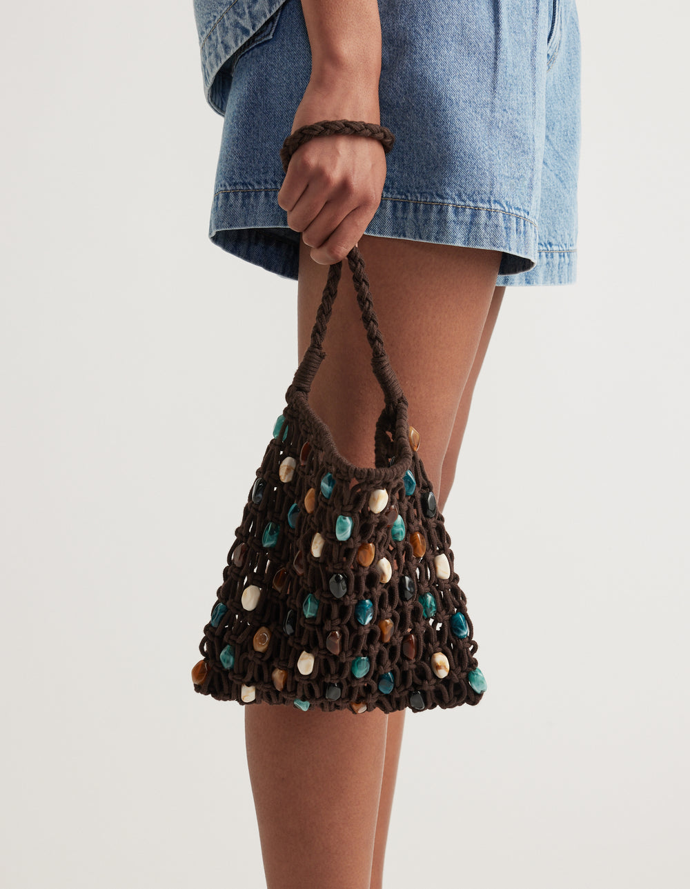 Woman carrying a handwoven brown handbag decorated with multicoloured gemstones and beads while wearing denim shorts