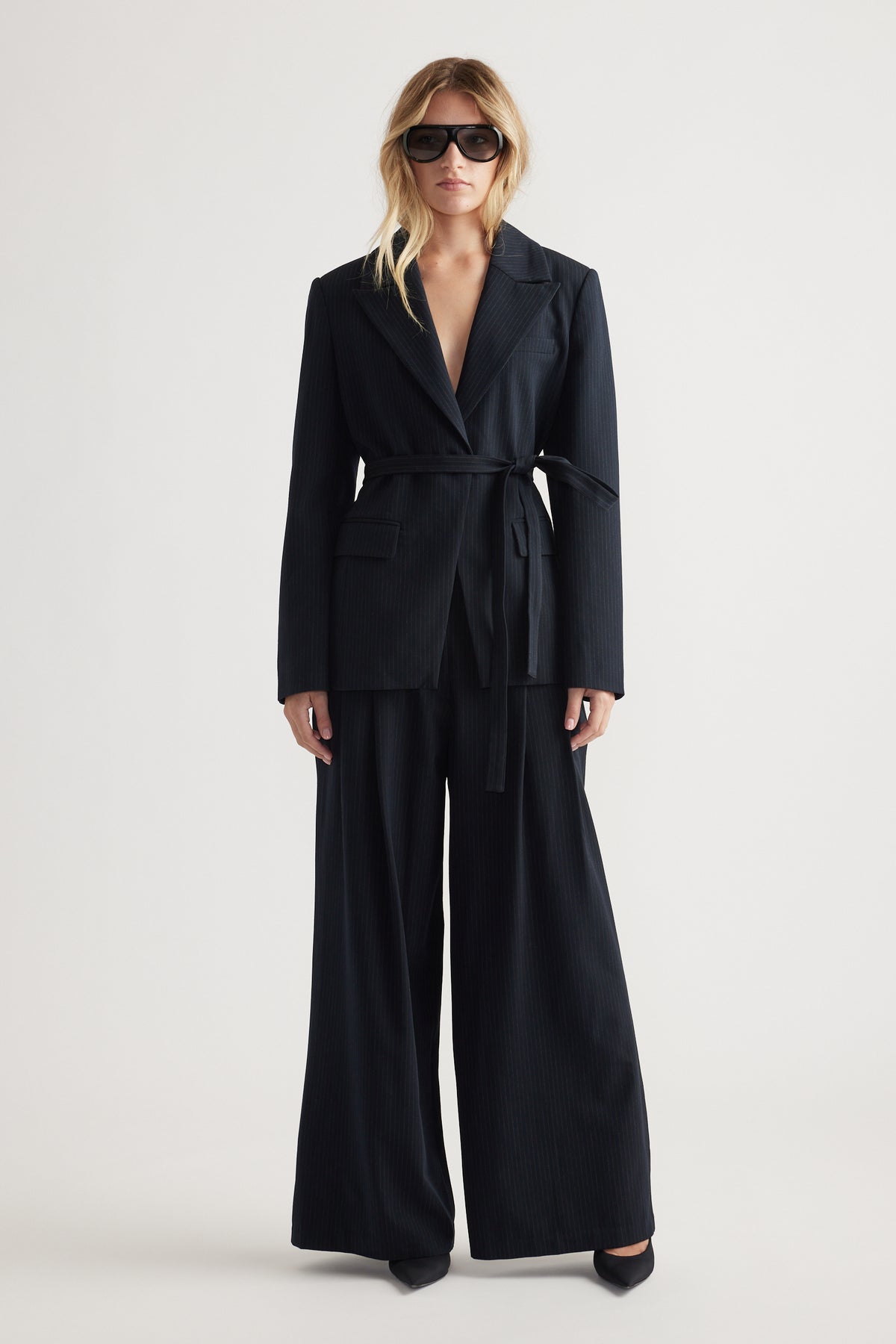 Woman wearing a navy tailored blazer with a belt, paired with wide-leg matching pants and oversized sunglasses