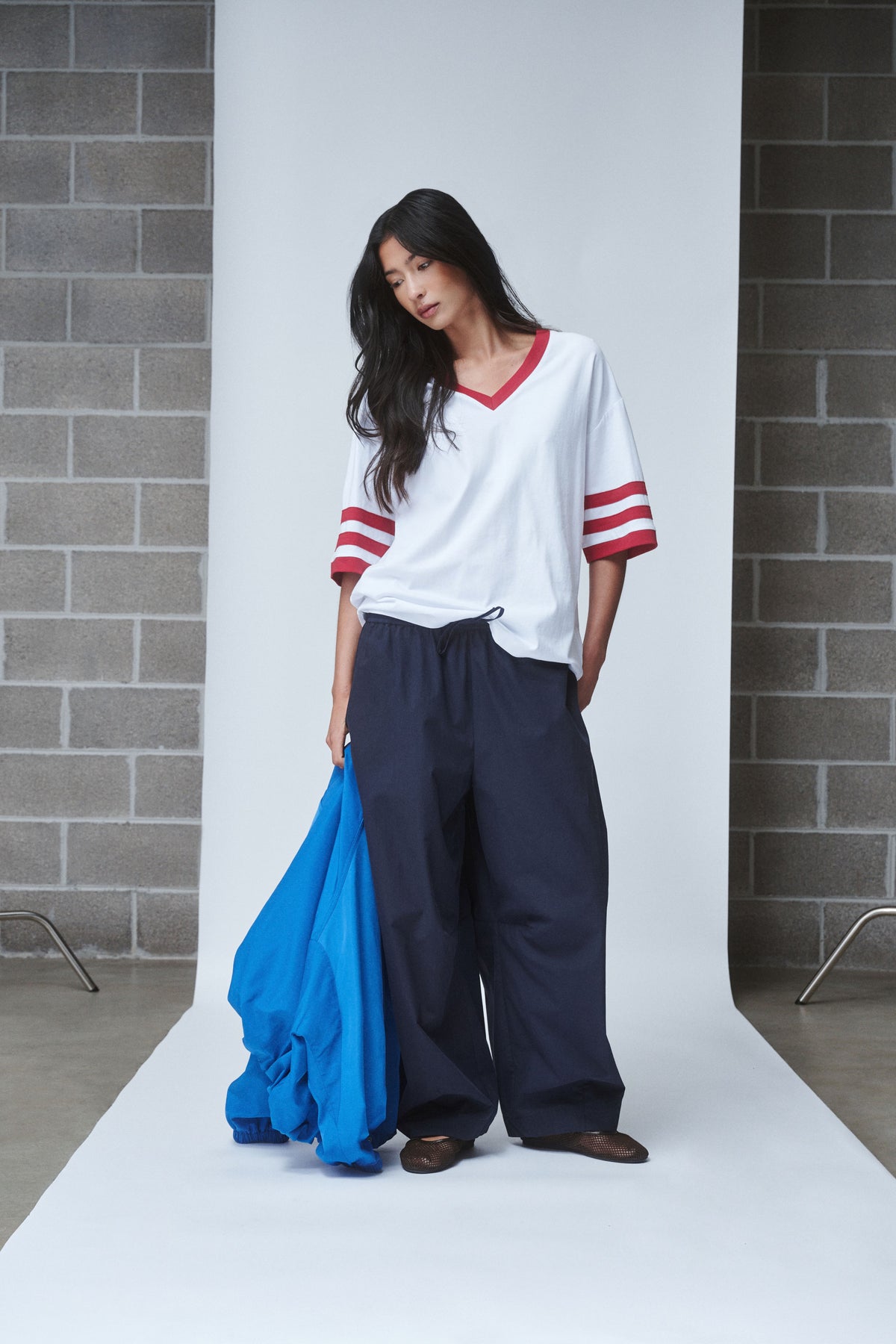 Woman wearing an oversized white V-neck tee with red striped sleeves and navy wide-leg pants, holding a blue garment