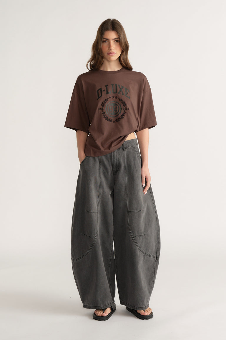 Model wearing a brown oversized graphic tee with dark grey barrel leg jeans and black sandals