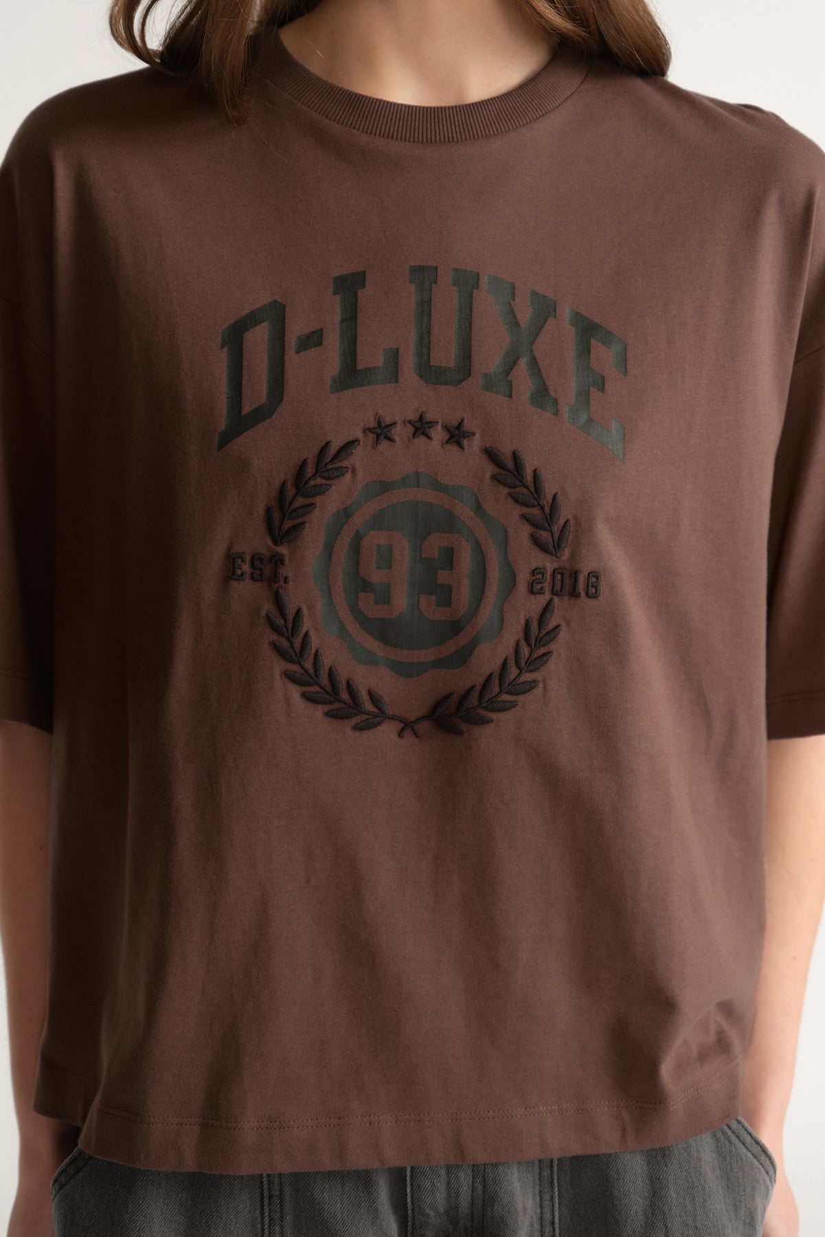 Woman wearing an oversized brown crest print tee with dark lettering and grey pants