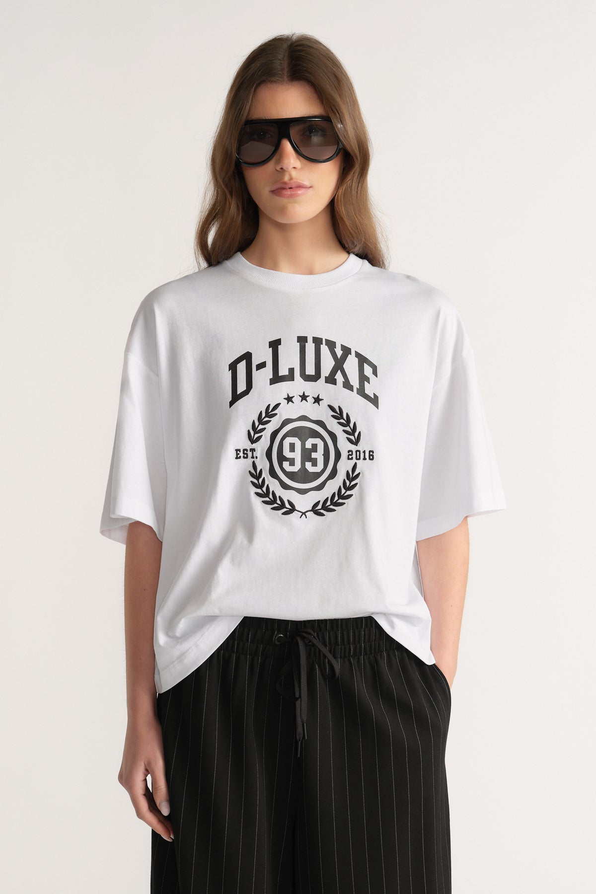 Model wearing an oversized white D-Luxe graphic tee with black pinstripe pants and sunglasses