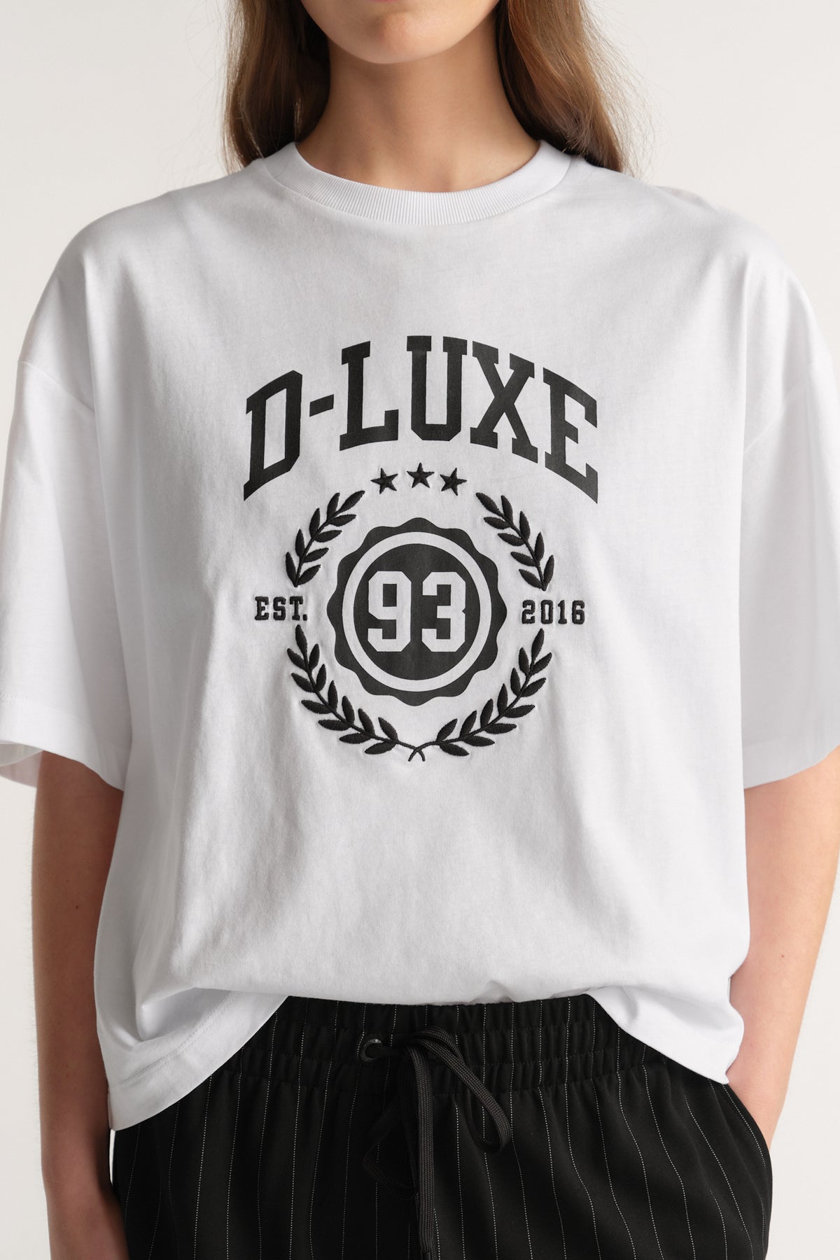 Model wearing a white oversized D-Luxe crest graphic tee with black pinstripe pants