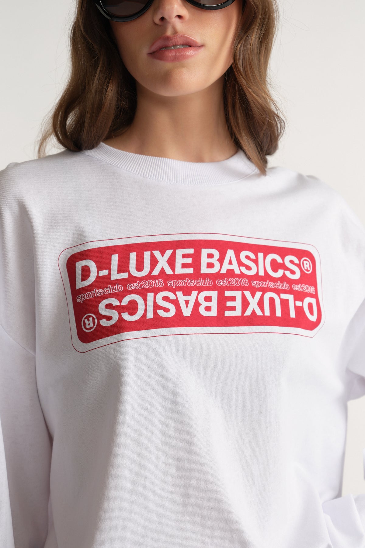 Woman wearing a white oversized long sleeve tee with a red D-Luxe Basics graphic прин?