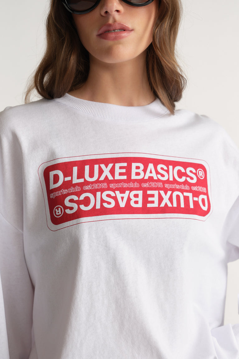 Woman wearing a white oversized long sleeve tee with a red D-Luxe Basics graphic прин?