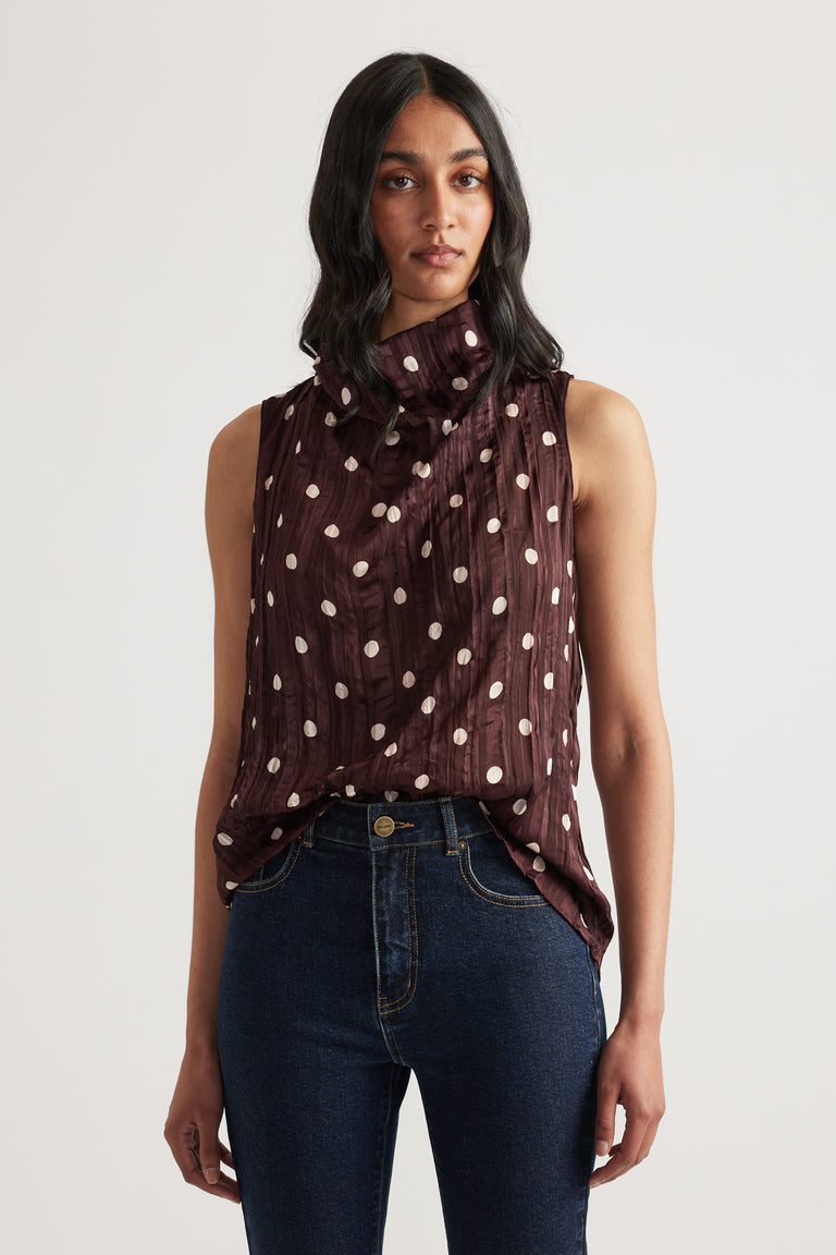 Woman wearing a burgundy textured sleeveless cowl neck top with polka dots and dark jeans