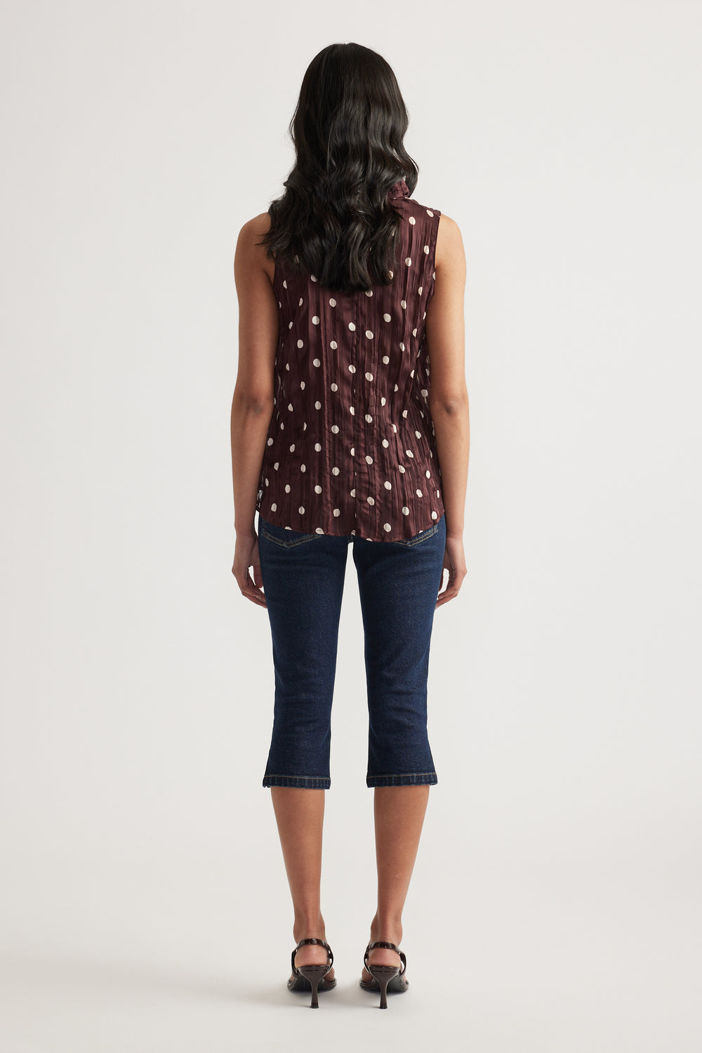 Woman seen from behind wearing a burgundy textured sleeveless top with a dotted pattern, cropped jeans and heels