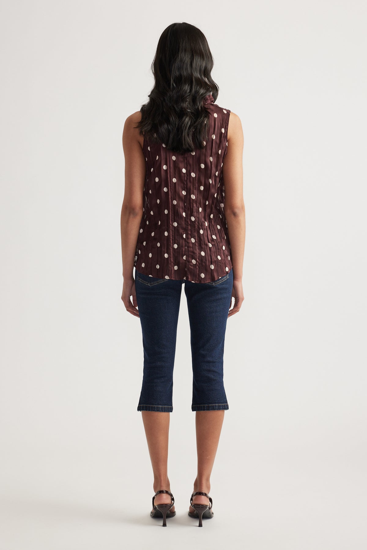 Woman seen from behind wearing a burgundy textured sleeveless top with a dotted pattern, cropped jeans and heels