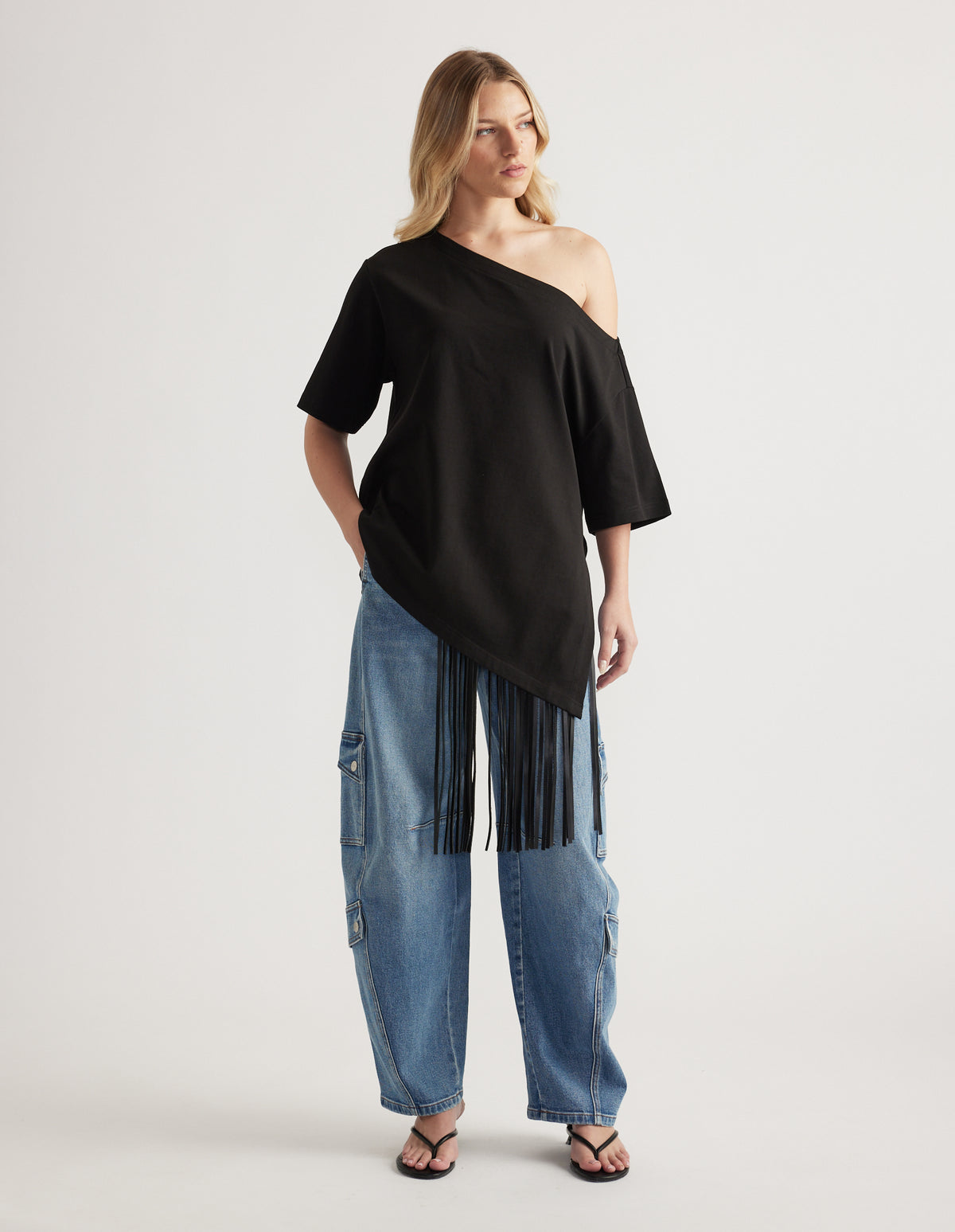 Woman wearing a black relaxed fit tee with asymmetric neckline and fringe hem, styled with cargo jeans and sandals
