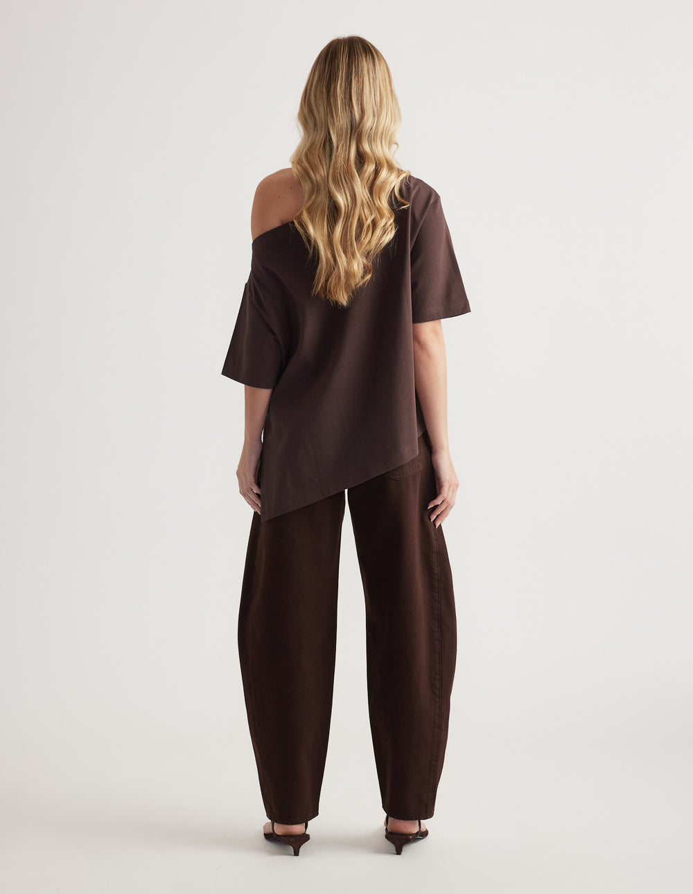 Back view of woman in a dark brown asymmetric tee with one shoulder exposed and matching relaxed pants