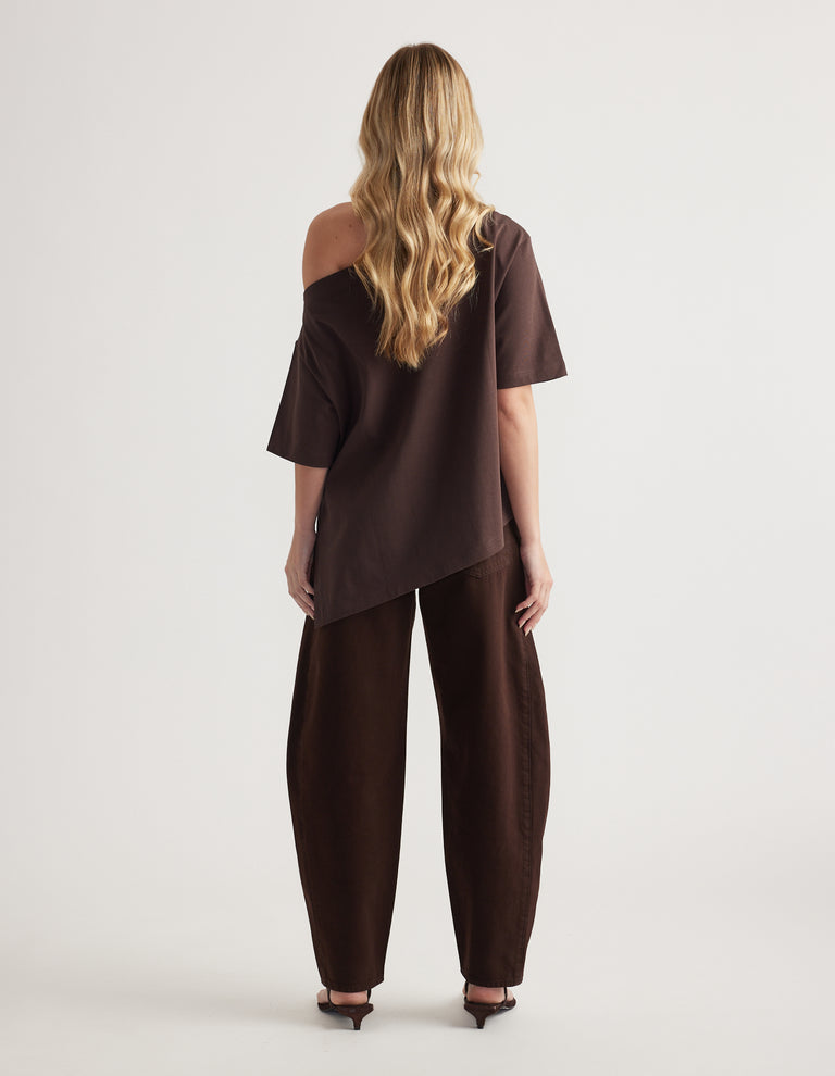 Back view of woman in a dark brown asymmetric tee with one shoulder exposed and matching relaxed pants