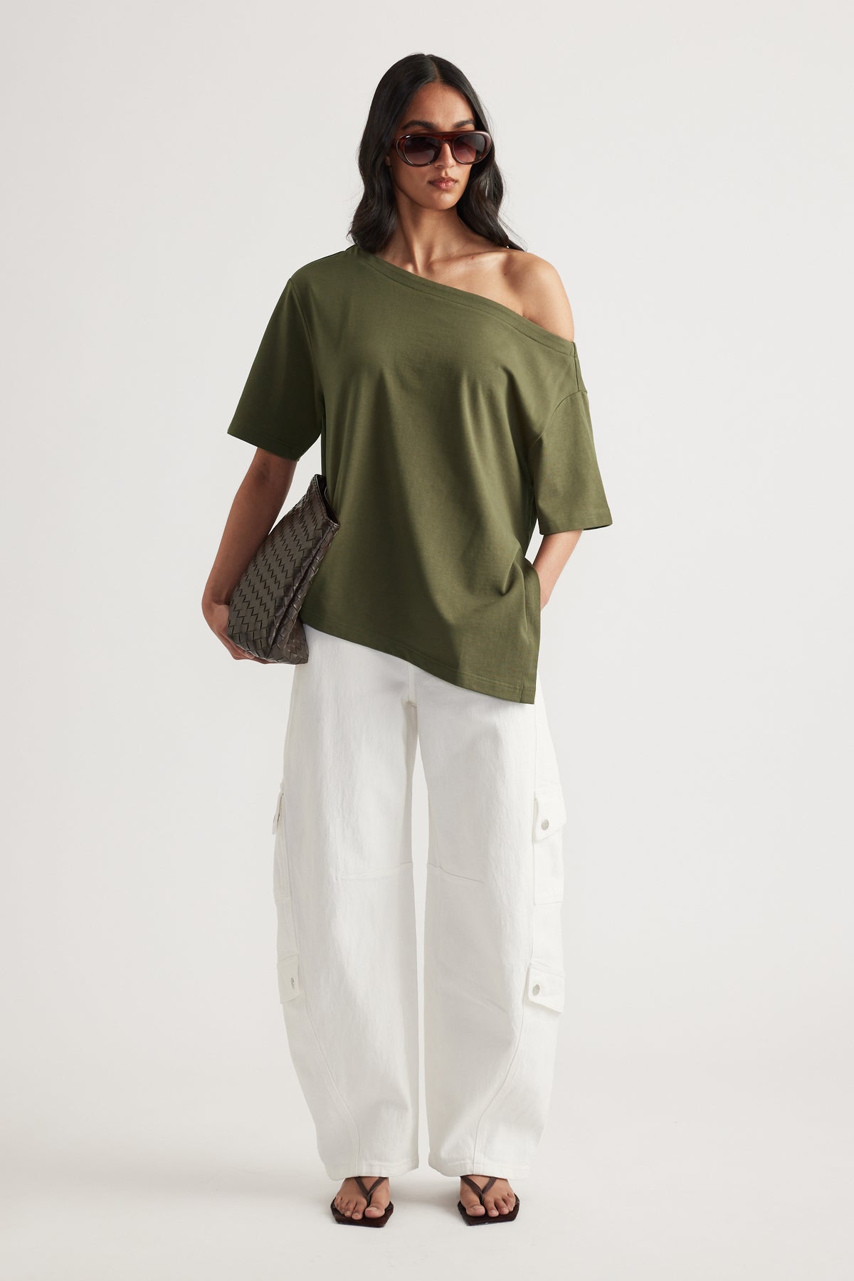 Woman styling an olive asymmetric off shoulder tee with white cargo pants, sunglasses, woven shoulder bag and sandals