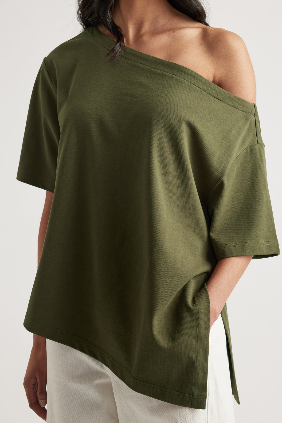 Woman wearing an olive green relaxed fit tee with an asymmetric neckline and hem, styled with white pants