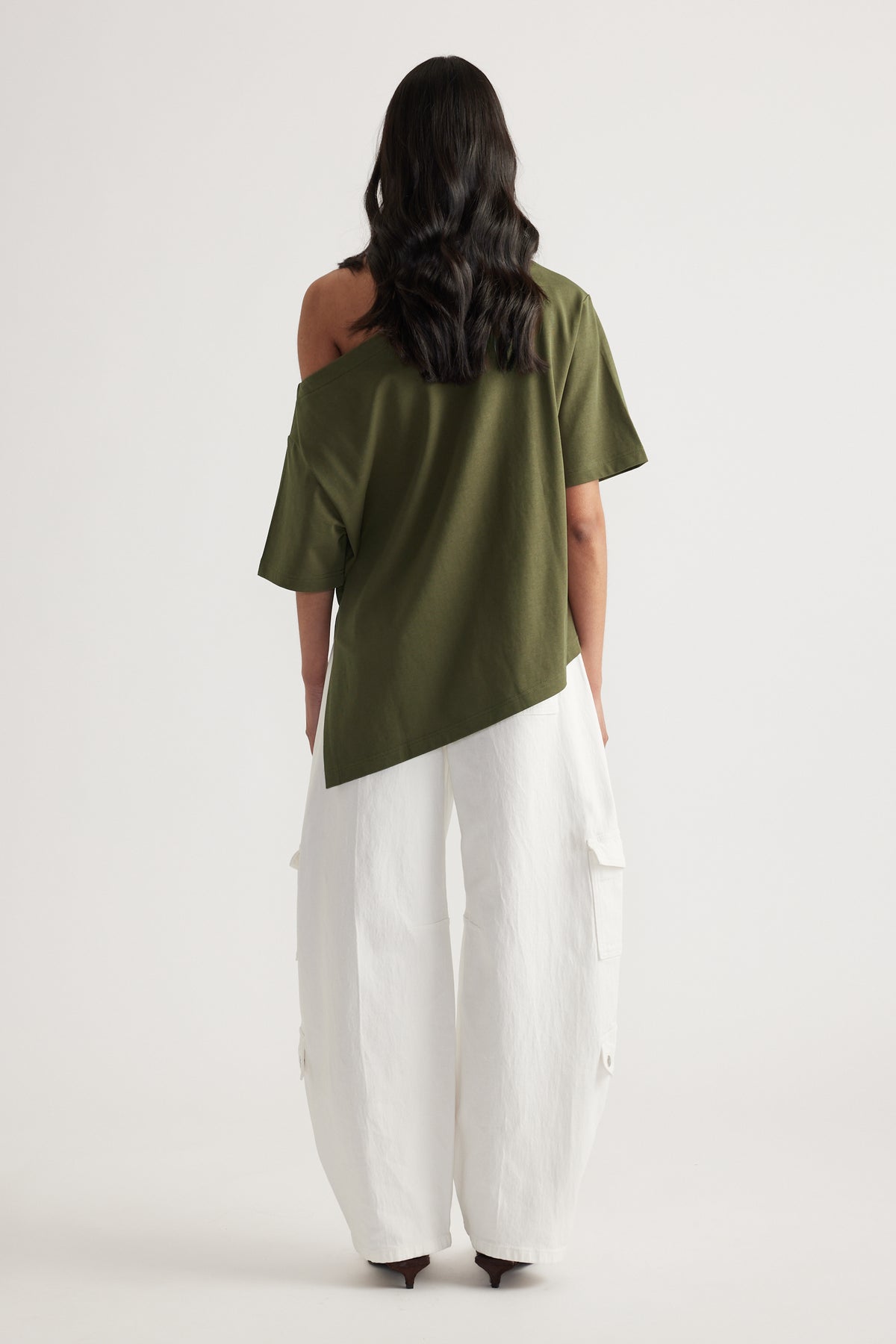 Back view of woman wearing an olive asymmetric tee with exposed shoulder, white cargo pants and heels