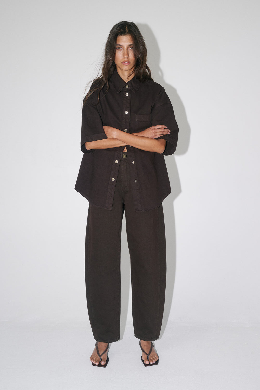 Woman wearing an oversized dark brown denim shirt with short sleeves, matching barrel leg pants and strappy sandals
