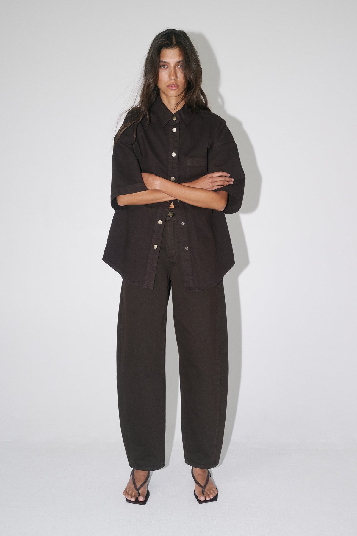 Woman wearing an oversized dark brown denim shirt with short sleeves, matching barrel leg pants and strappy sandals