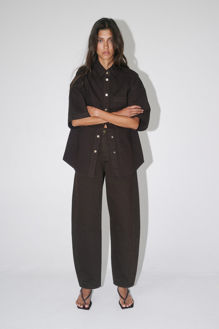 Woman wearing an oversized dark brown denim shirt with short sleeves, matching barrel leg pants and strappy sandals