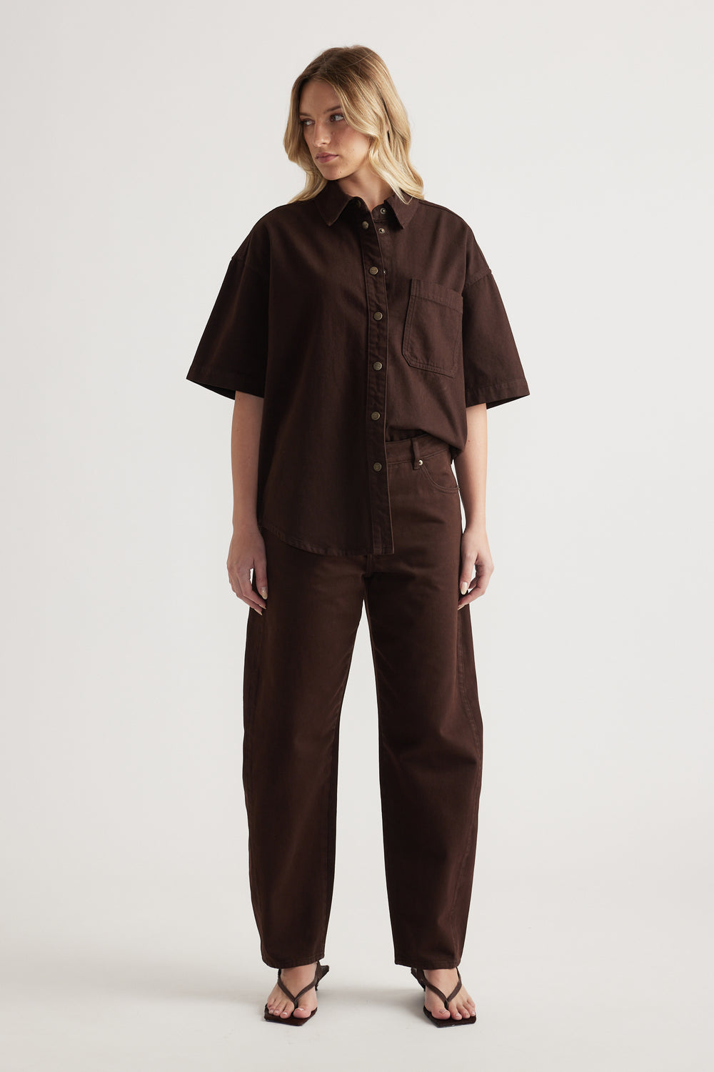 Woman wearing an oversized brown denim shirt with matching barrel leg jeans and strappy flat sandals