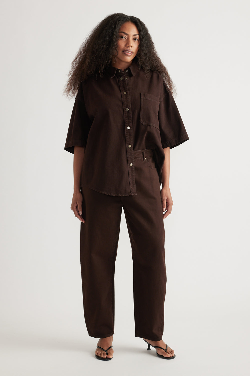 Woman wearing an oversized brown denim shirt with short sleeves, matching barrel-leg jeans and sandals