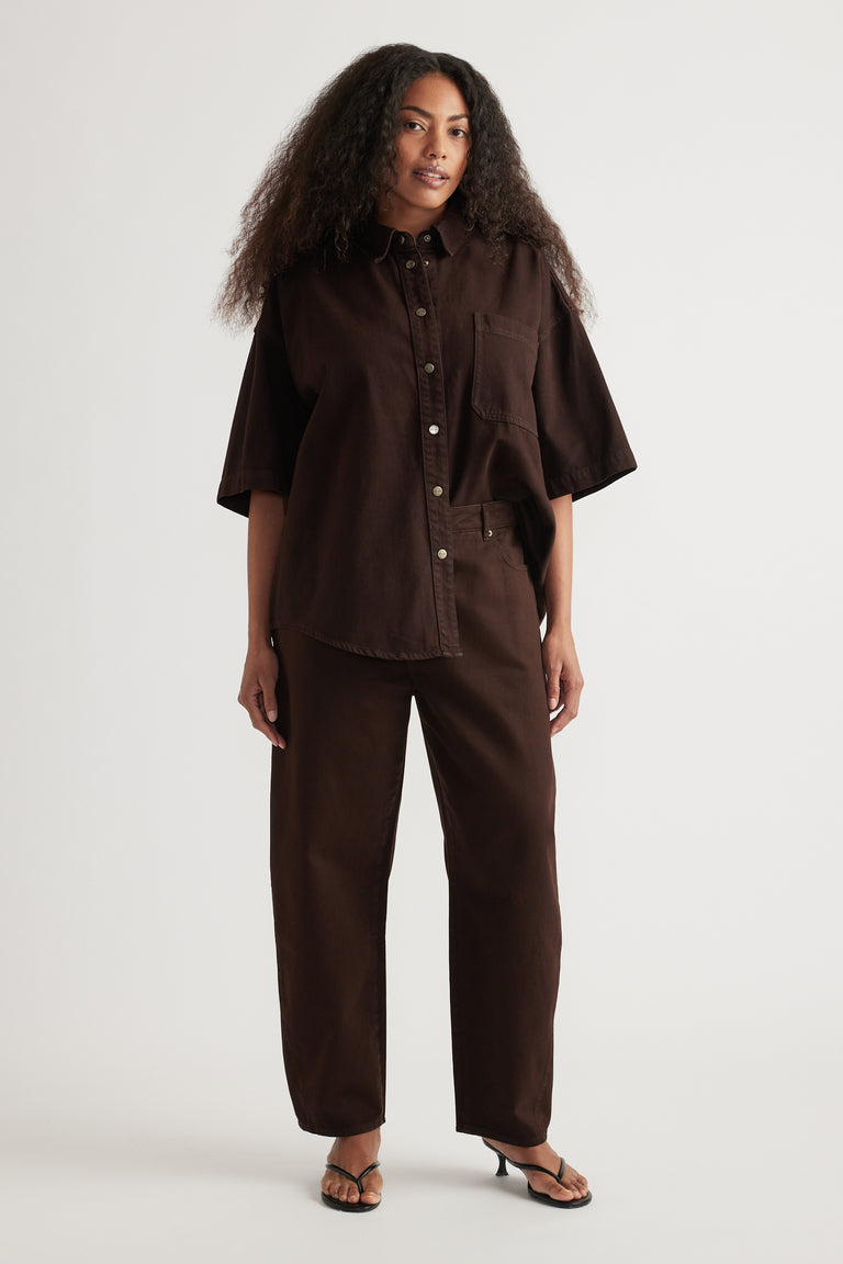 Woman wearing an oversized brown denim shirt with short sleeves, matching barrel-leg jeans and sandals