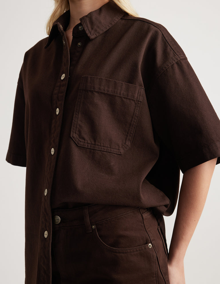 Model wearing an oversized brown denim shirt with short sleeves, collar and chest pocket