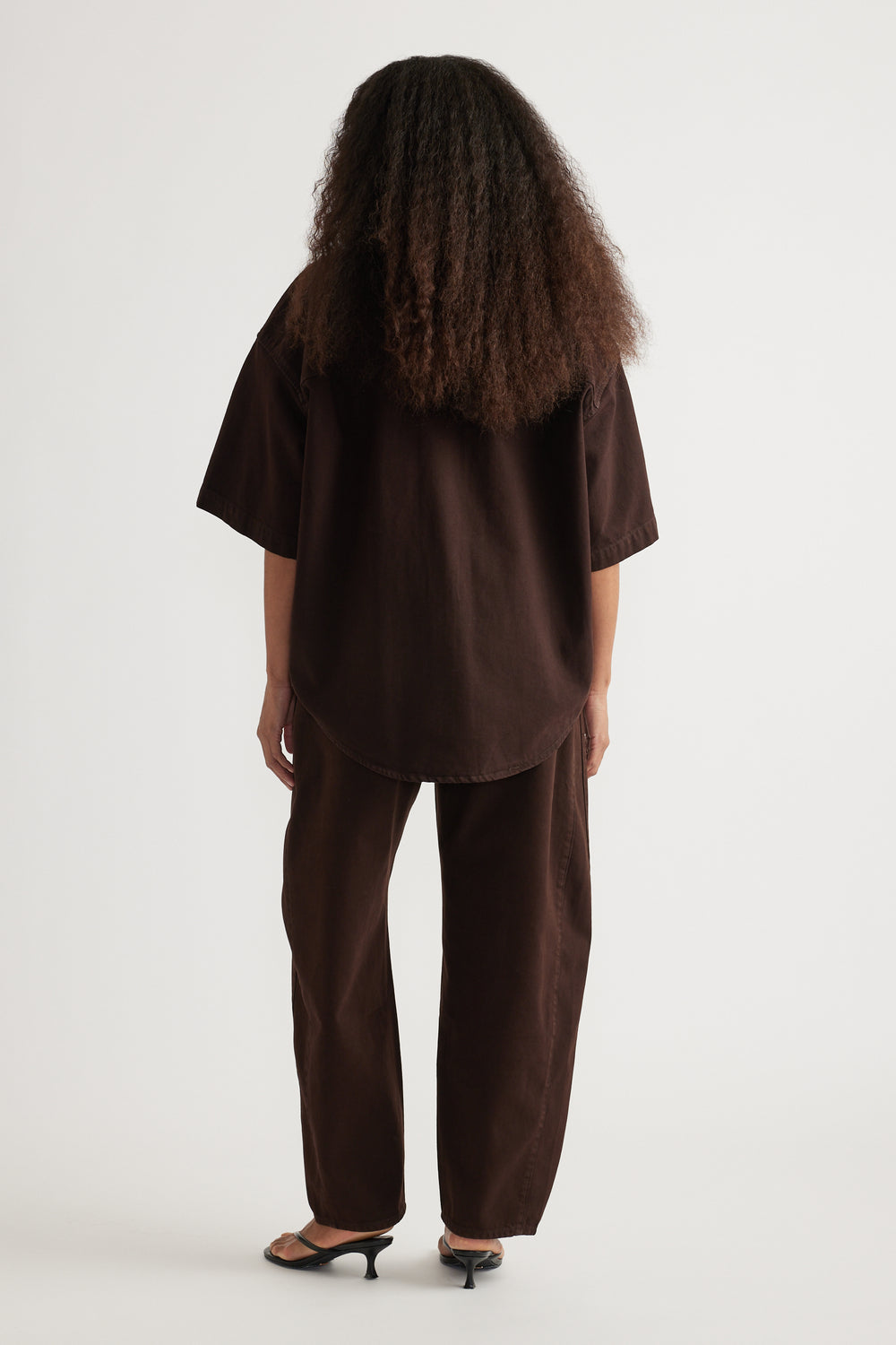 Back view of a model in a brown oversized denim shirt with relaxed matching pants and heeled sandals