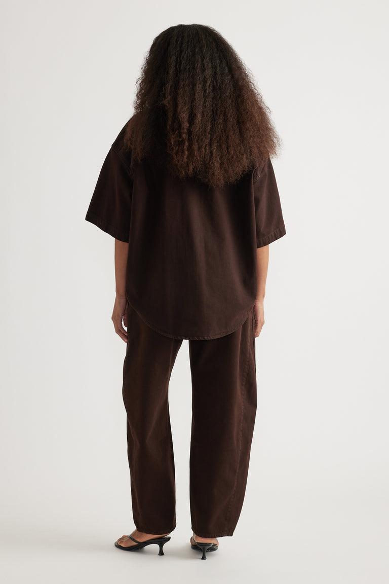 Back view of a model in a brown oversized denim shirt with relaxed matching pants and heeled sandals
