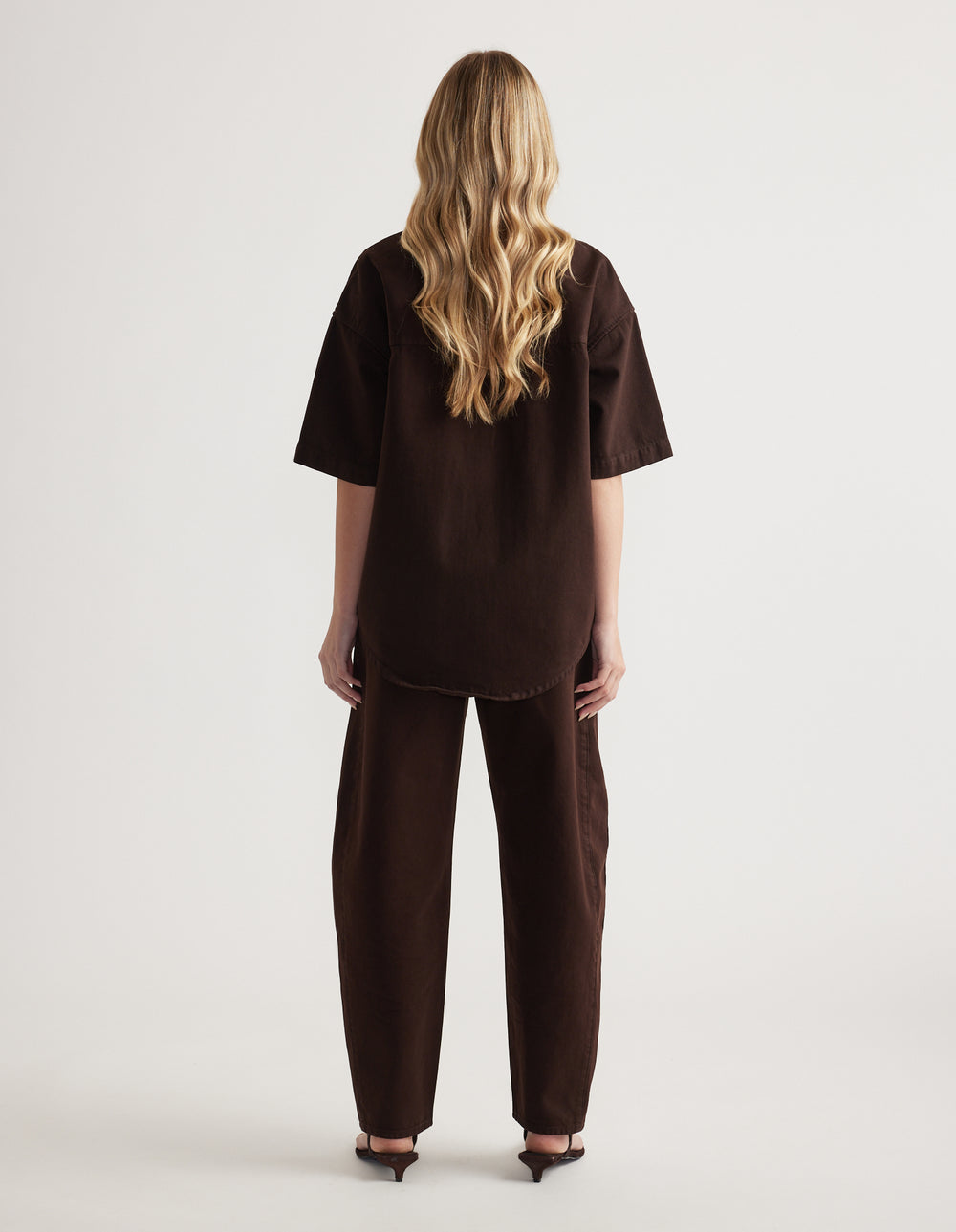 Back view of a woman wearing an oversized brown denim shirt with short sleeves and matching barrel-leg jeans