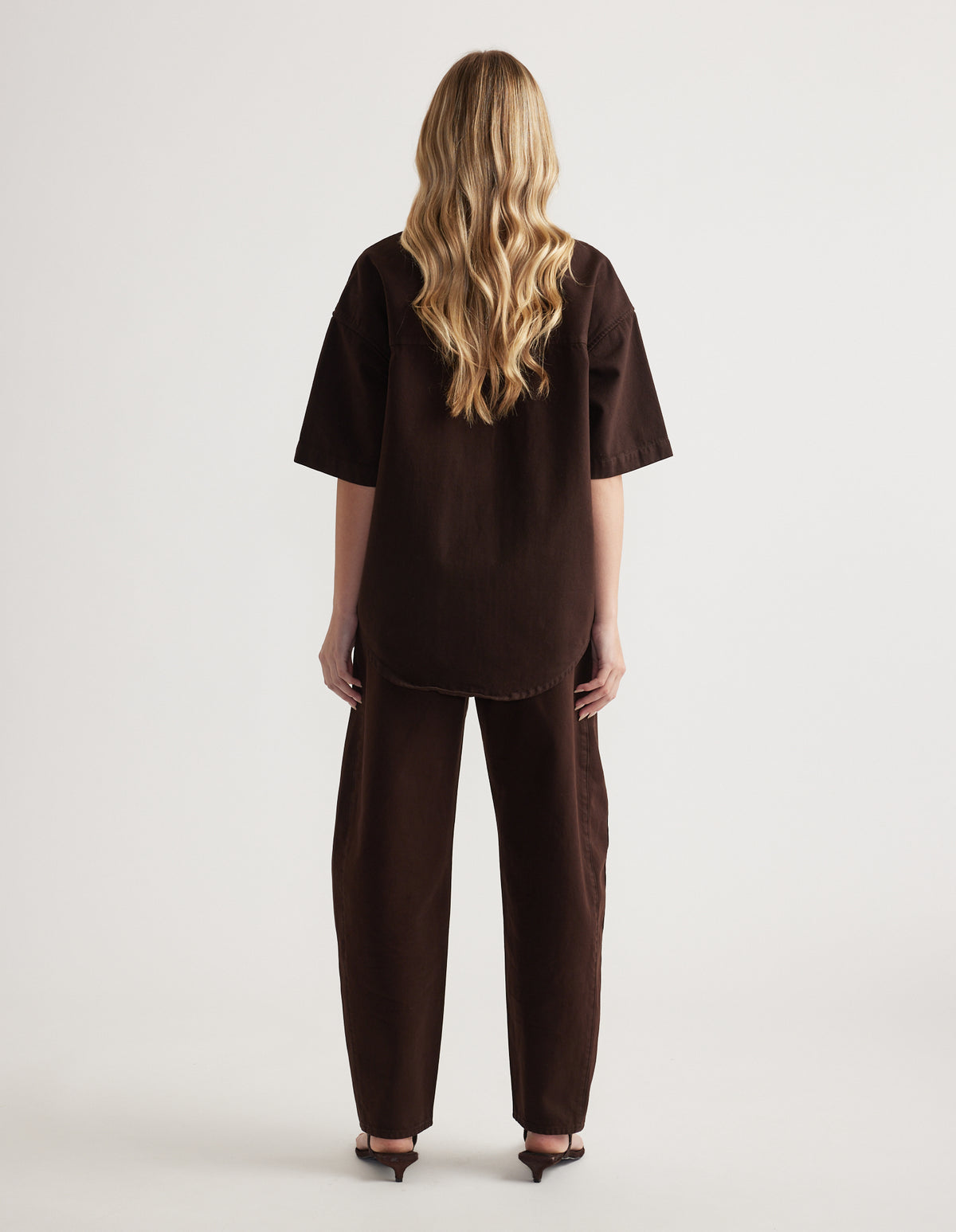 Back view of a woman wearing an oversized brown denim shirt with short sleeves and matching barrel-leg jeans