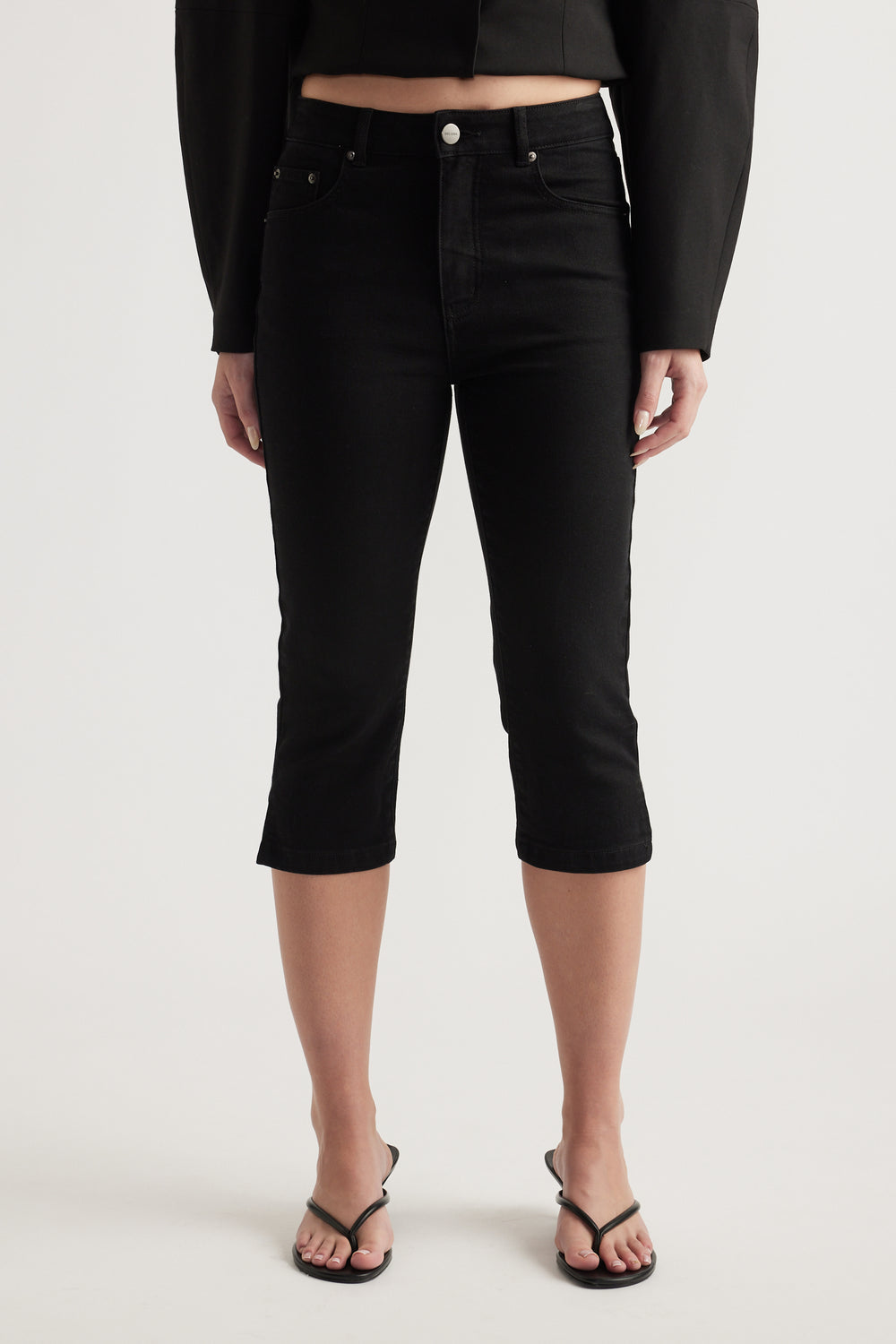 Woman wearing high rise black denim capri jeans with a cropped long sleeve top and flat sandals