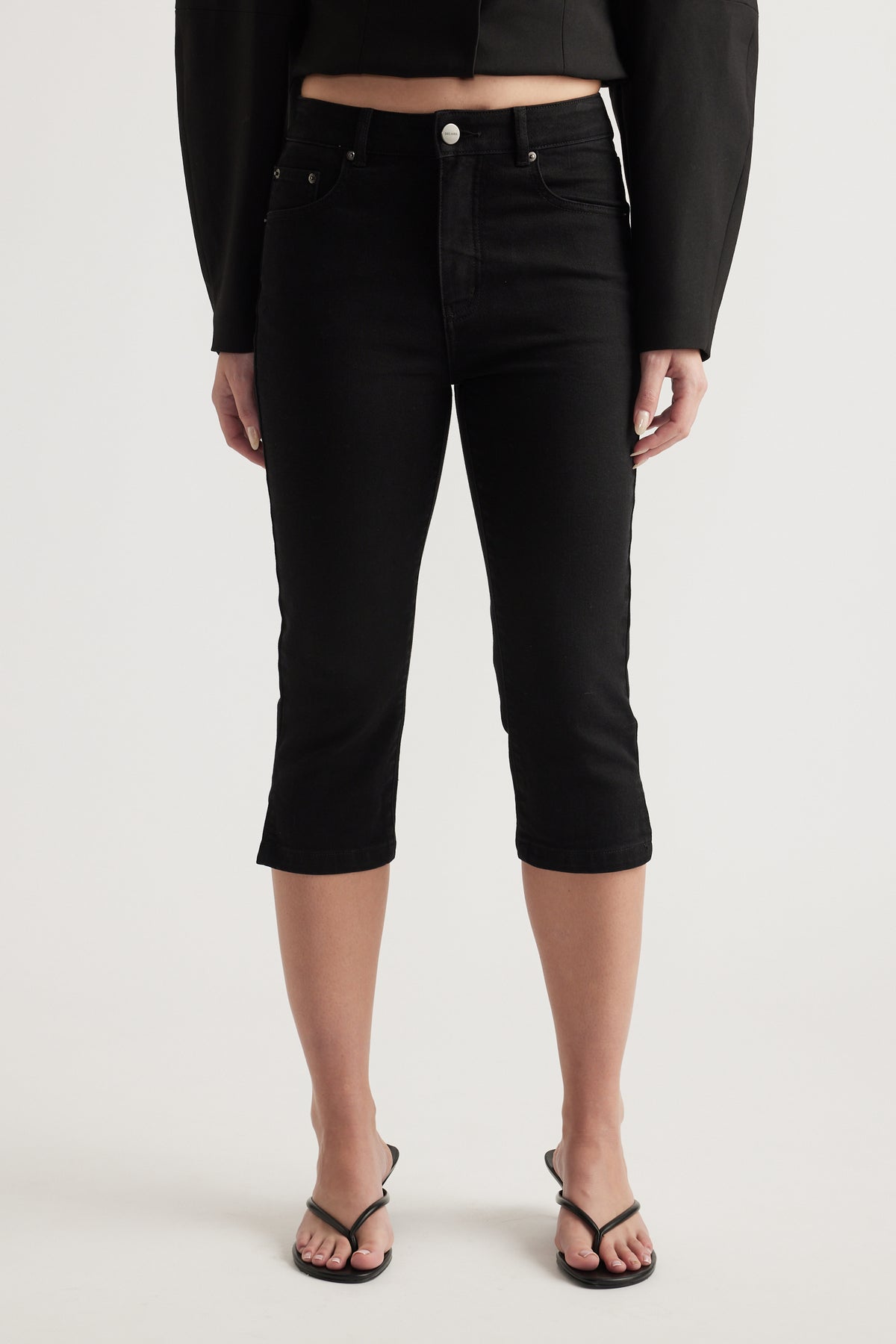 Woman wearing high rise black denim capri jeans with a cropped long sleeve top and flat sandals