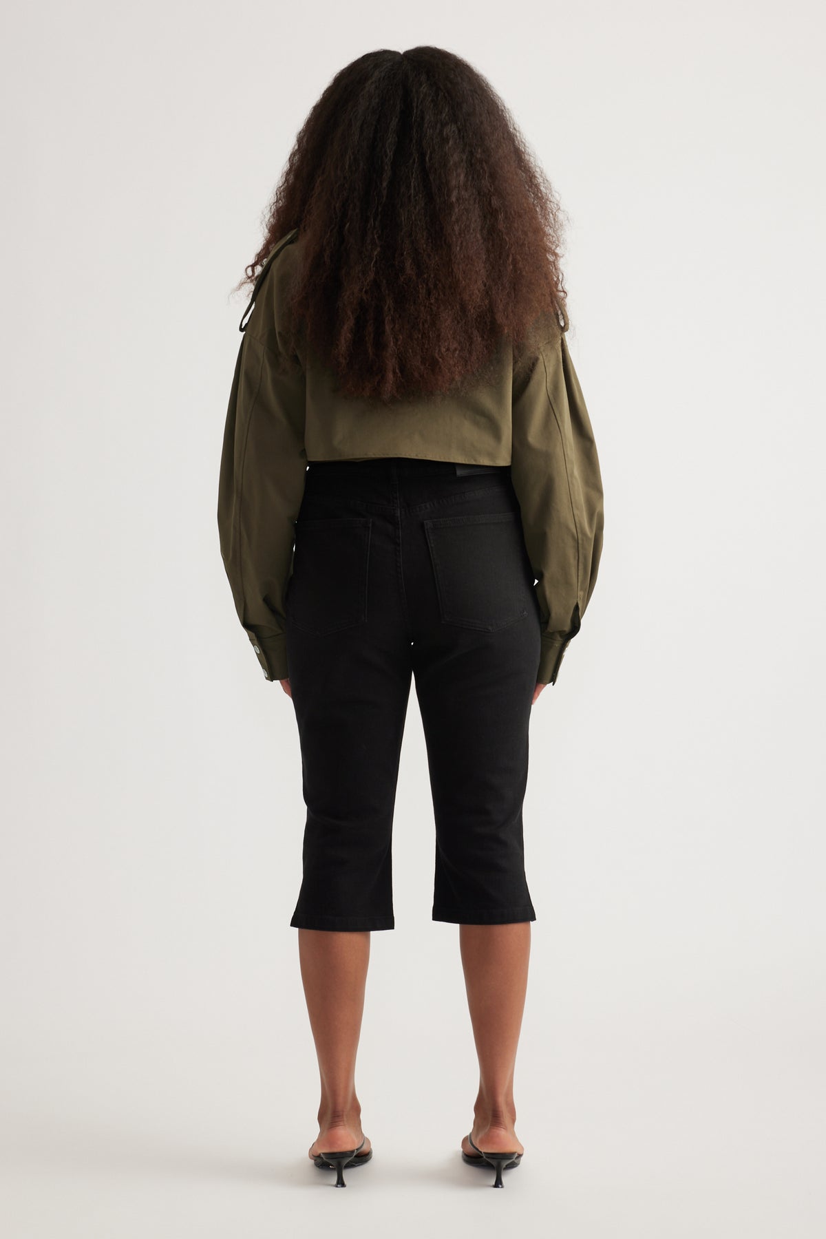 Back view of a model wearing black high-rise denim capris with an olive cropped jacket and black heels
