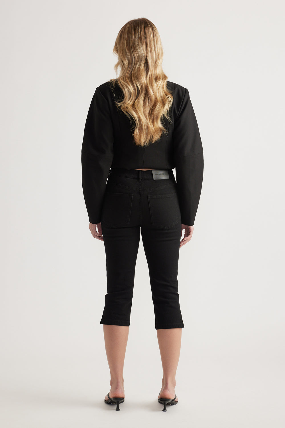 Woman viewed from behind wearing a black top with black denim capri pants and heels