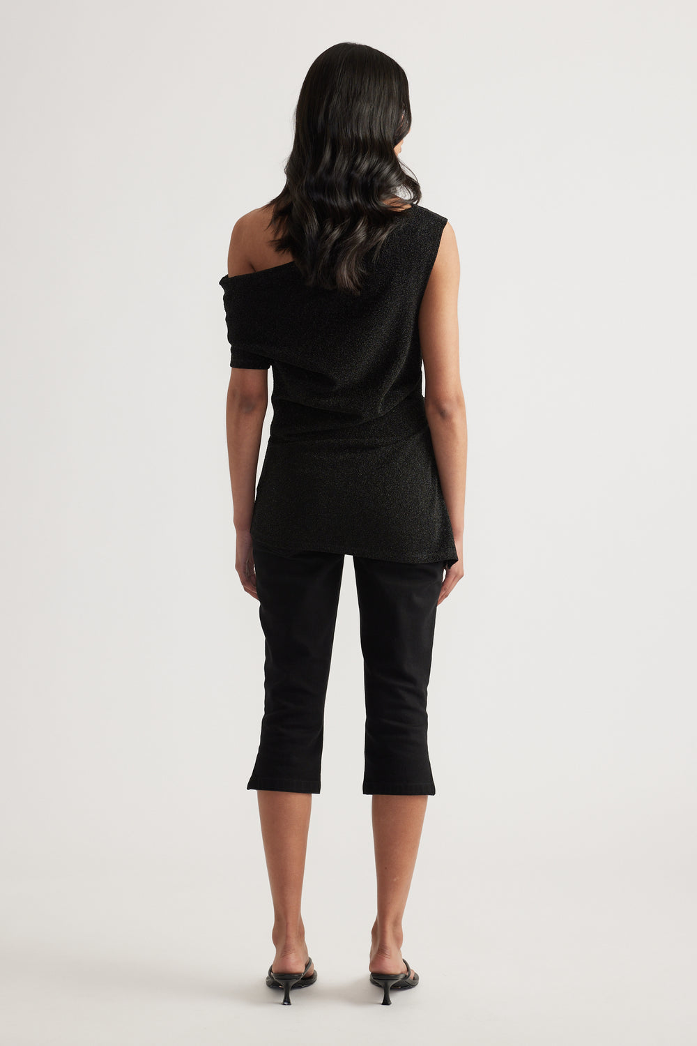 Back view of a woman in a black asymmetric off shoulder top, cropped pants and heels