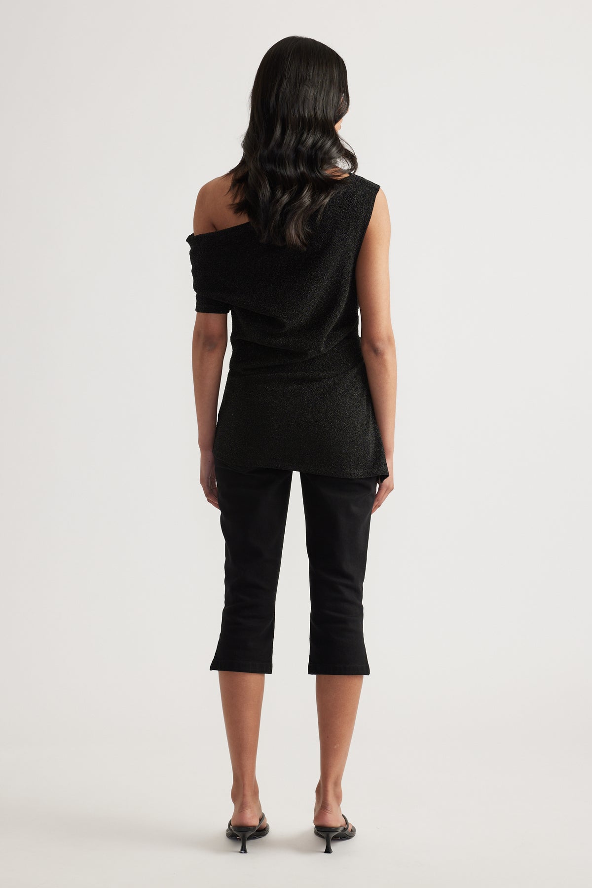 Back view of a woman in a black asymmetric off shoulder top, cropped pants and heels