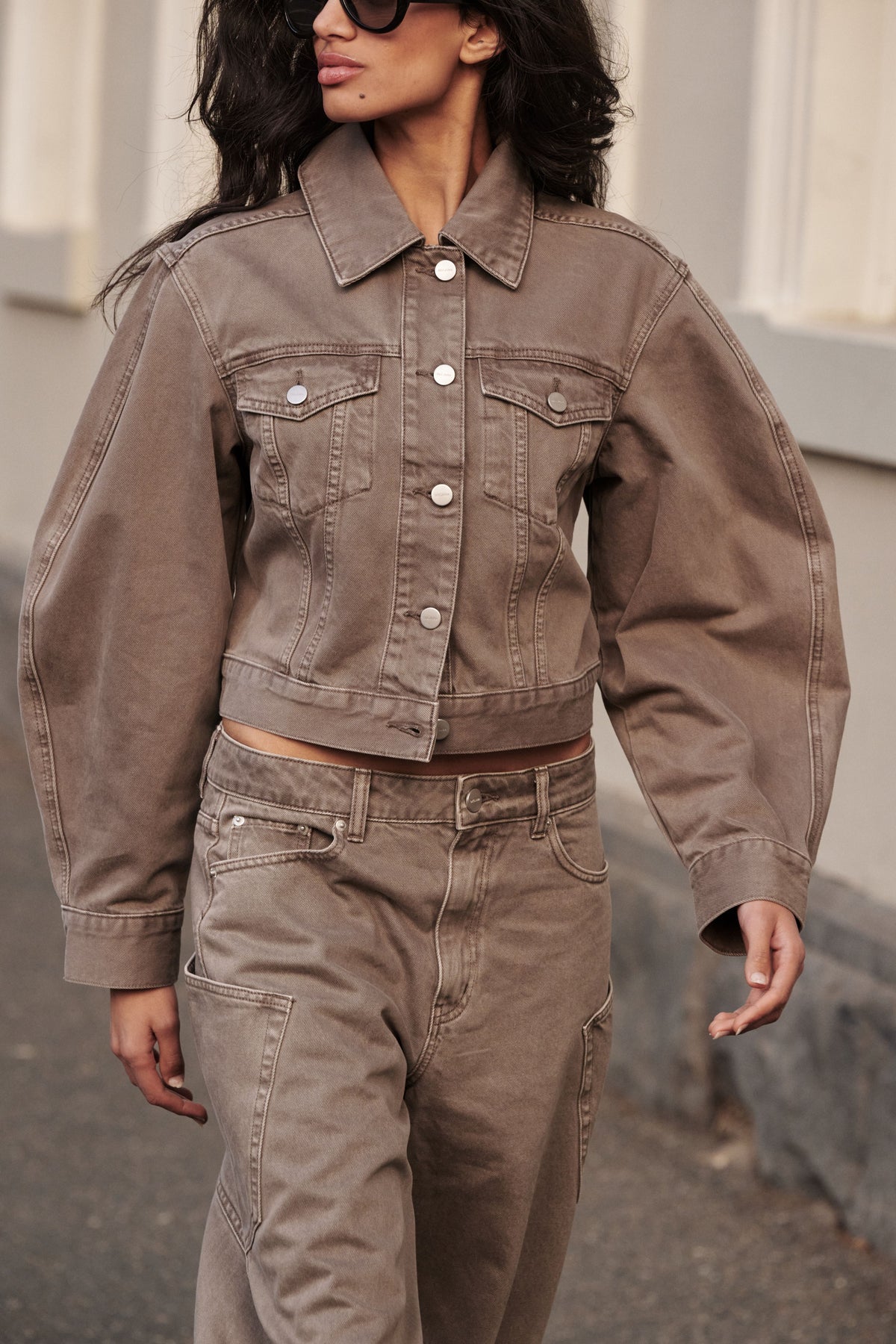 Woman wearing a brown cropped denim jacket with long barrel sleeves, chest pockets and a button front