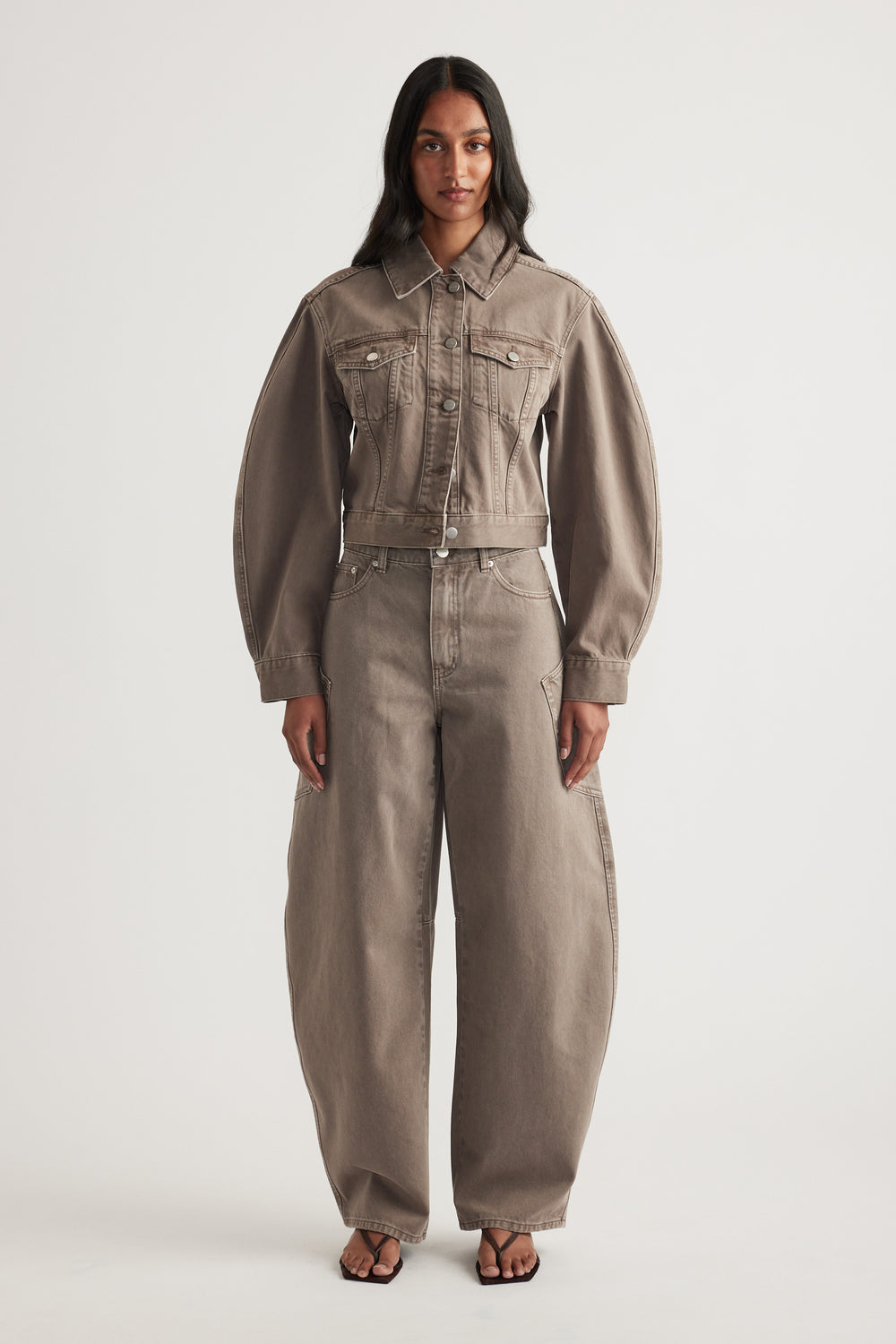 Model in a cropped taupe denim jacket with chest pockets, matching barrel leg jeans and black sandals